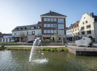 Stadtplatz mit einem Brunnen im Vordergrund, umgeben von mehrstöckigen Gebäuden und Geschäften, bei klarem Himmel.
